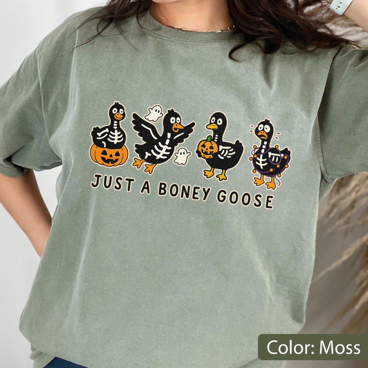 May include: Moss green t-shirt with a Halloween graphic. The graphic features four cartoon geese with skeleton designs, pumpkins, and ghosts. The text "JUST A BONEY GOOSE" is printed below the geese.