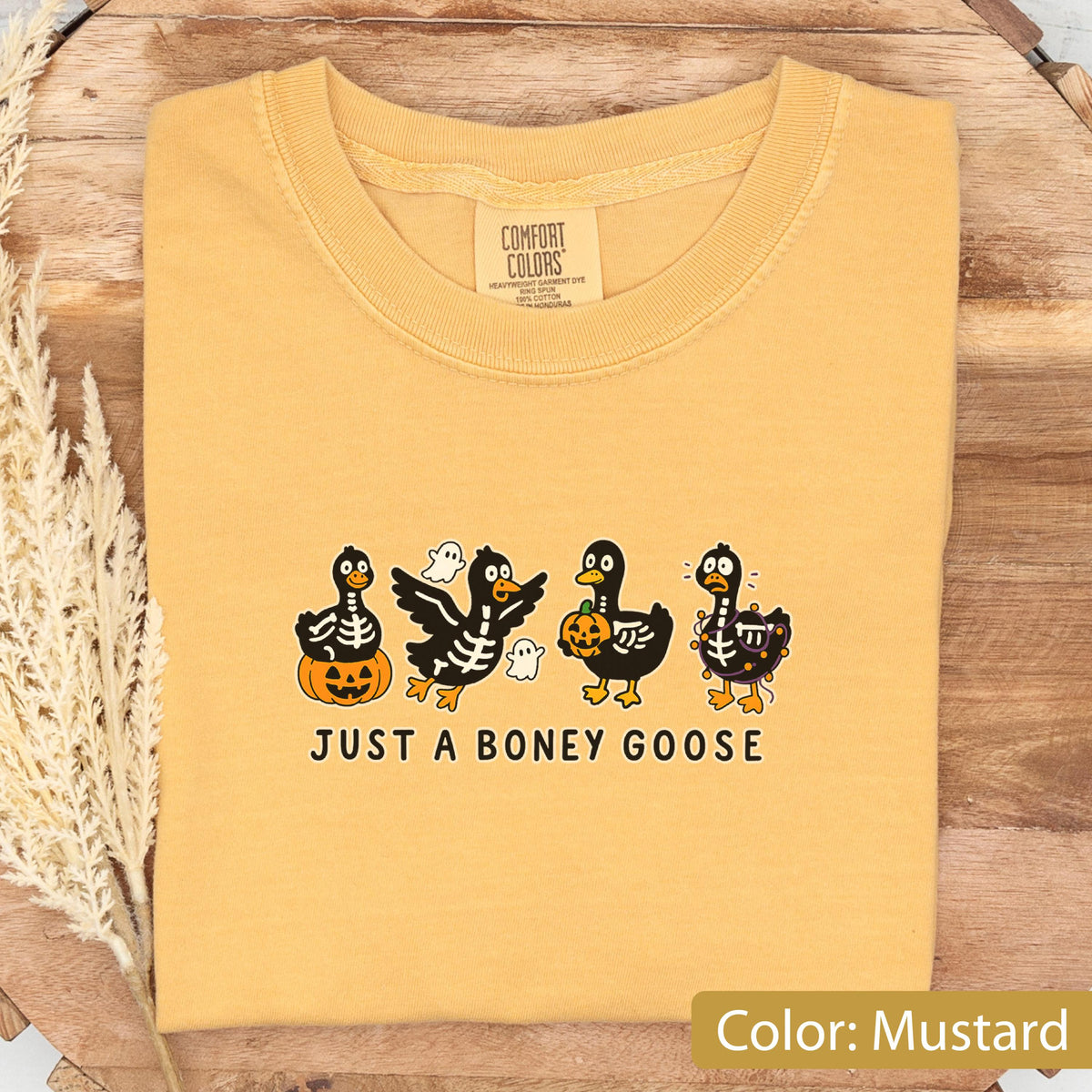 May include: Mustard-colored t-shirt with a crew neck. The shirt features a graphic of four cartoon geese in Halloween costumes, with the text "JUST A BONEY GOOSE" below. The geese are decorated with skeletons, pumpkins, and ghosts.