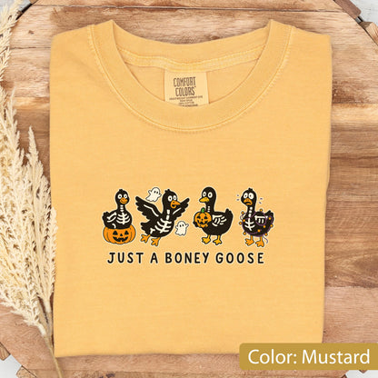 May include: Mustard-colored t-shirt with a crew neck. The shirt features a graphic of four cartoon geese in Halloween costumes, with the text "JUST A BONEY GOOSE" below. The geese are decorated with skeletons, pumpkins, and ghosts.