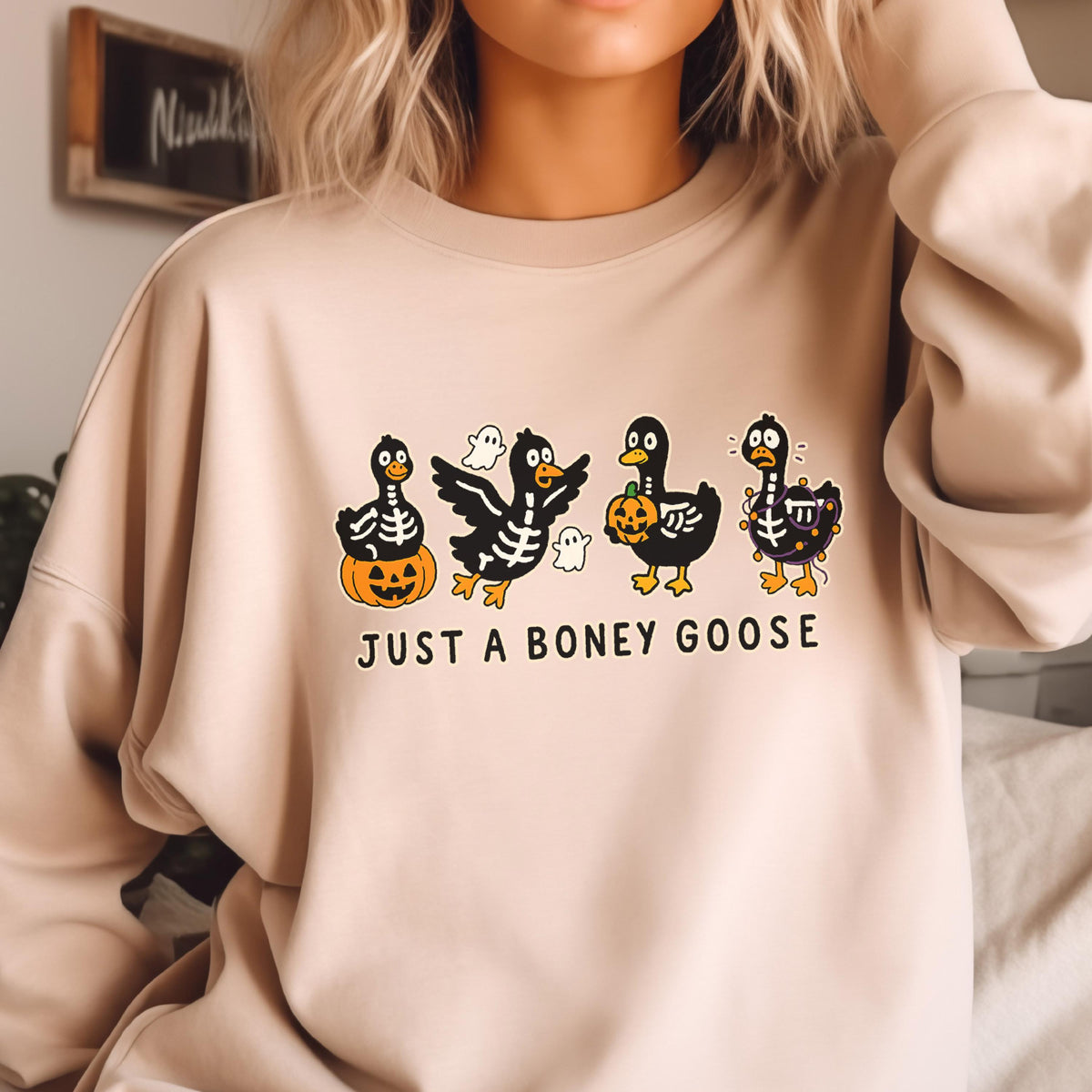 May include: Beige sweatshirt featuring a Halloween-themed graphic. The design includes four cartoon geese dressed in skeleton costumes, holding pumpkins and surrounded by ghosts. The text "JUST A BONEY GOOSE" is printed below the geese.