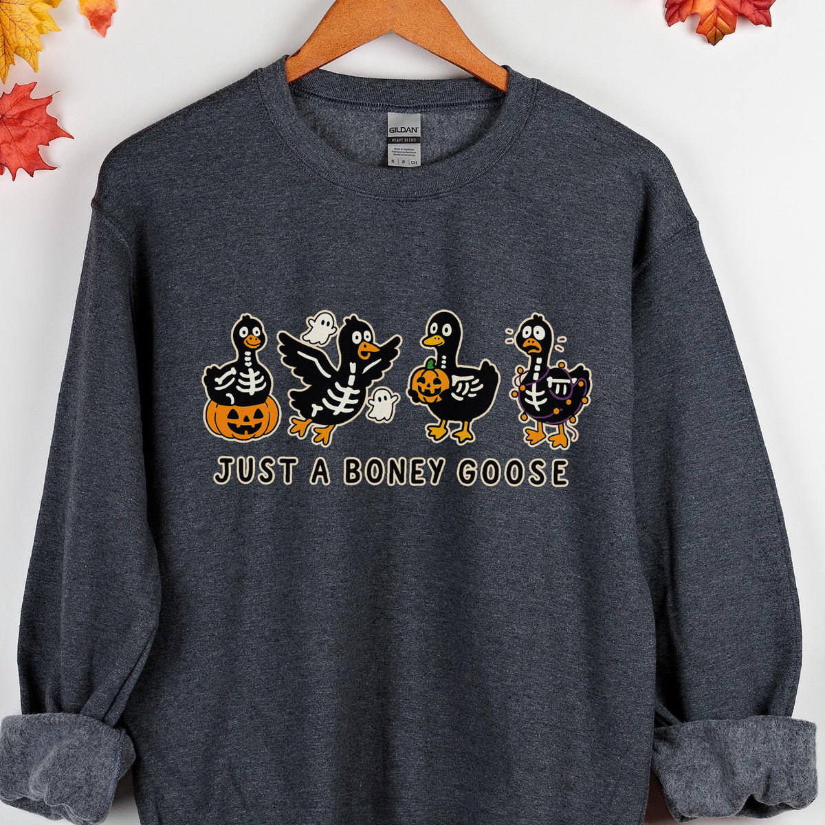 May include: Dark gray sweatshirt featuring a Halloween-themed graphic. The design includes four cartoon geese with skeleton and pumpkin details, along with the text "JUST A BONEY GOOSE". The sweatshirt is hanging on a wooden hanger.