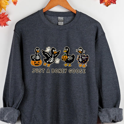 May include: Dark gray sweatshirt featuring a Halloween-themed graphic. The design includes four cartoon geese with skeleton and pumpkin details, along with the text "JUST A BONEY GOOSE". The sweatshirt is hanging on a wooden hanger.