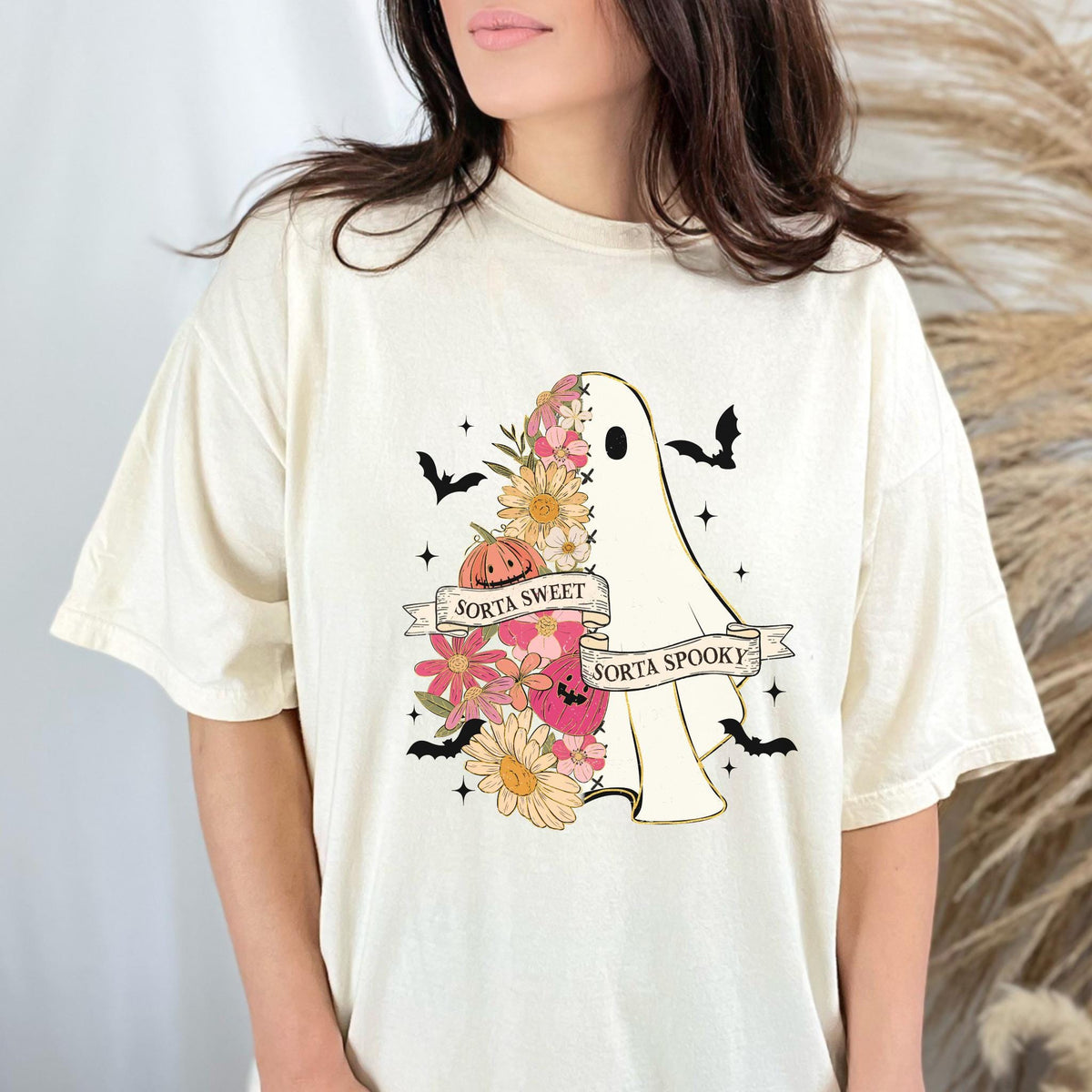 May include: Cream-colored t-shirt featuring a ghost design with floral accents, a pumpkin, and bats. The ghost has the text "Sorta Sweet Sorta Spooky" on a banner. A fun, festive design for Halloween.