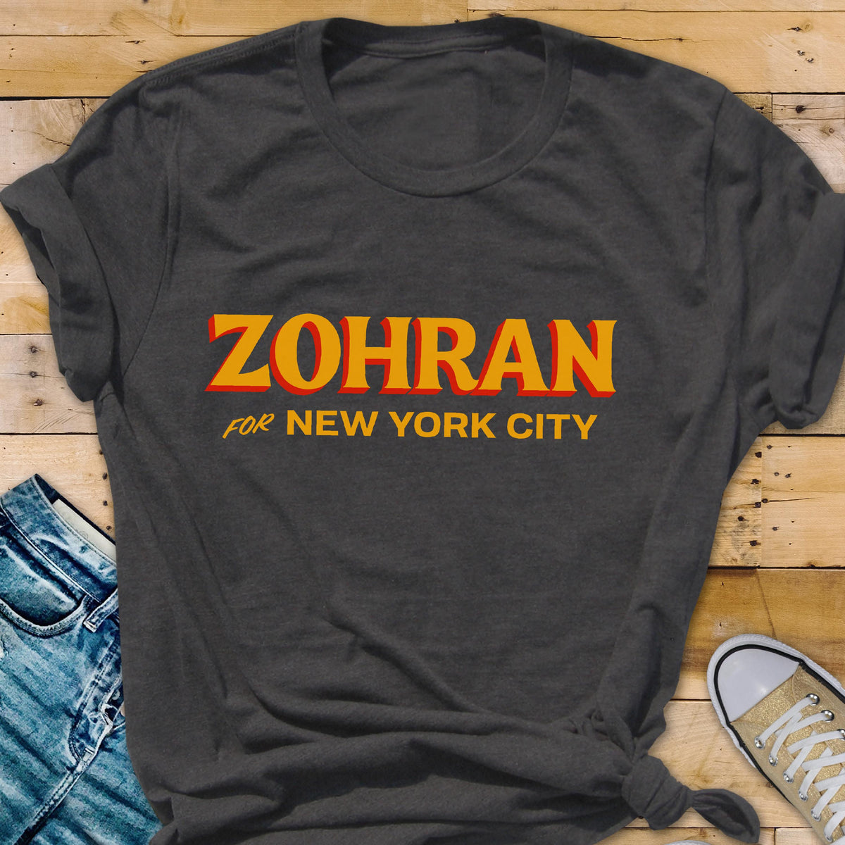 May include: A dark charcoal gray short-sleeved t-shirt featuring the text ZOHRAN FOR NEW YORK CITY printed in bold yellow letters with a red drop shadow effect. The shirt has a crew neckline and is made of a soft, textured fabric material.