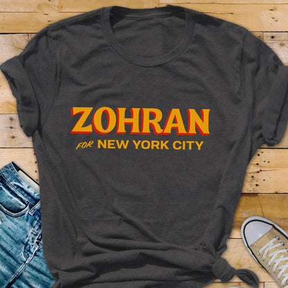 May include: A dark charcoal gray short-sleeved t-shirt featuring the text ZOHRAN FOR NEW YORK CITY printed in bold yellow letters with a red drop shadow effect. The shirt has a crew neckline and is made of a soft, textured fabric material.
