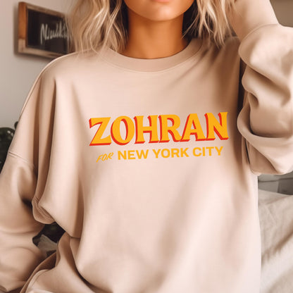 May include: A beige sweatshirt with the word "ZOHRAN" in orange and yellow block letters with a red shadow. Below the word is the phrase "FOR NEW YORK CITY" in smaller orange letters. The sweatshirt is a neutral color.