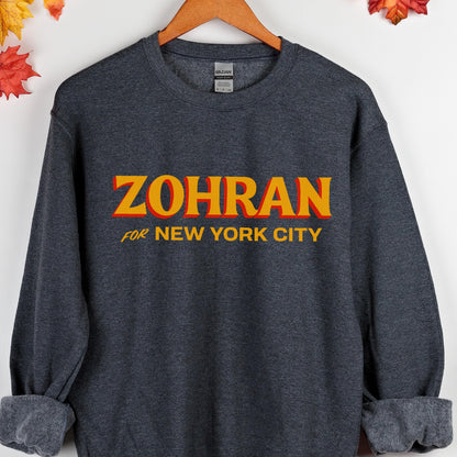 May include: A dark gray sweatshirt with the word "ZOHRAN" in large, orange and yellow letters across the chest. Below the word "ZOHRAN" is the text "FOR NEW YORK CITY" in smaller, white letters. The sweatshirt is hanging on a wooden hanger.