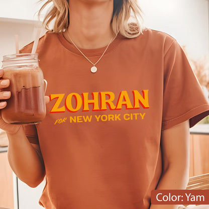 May include: A yam-colored t-shirt with the text "ZOHRAN" in yellow and orange, with "FOR NEW YORK CITY" below. The shirt is short-sleeved and has a round neckline. A person is wearing the shirt and holding a glass of a brown beverage.