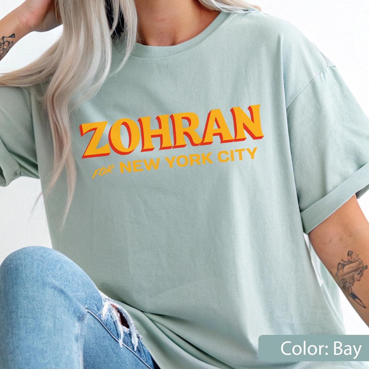 May include: A light blue t-shirt with the text "ZOHRAN" in orange letters with a red outline, and "FOR NEW YORK CITY" in smaller orange letters. The shirt is a solid color and has rolled-up sleeves.