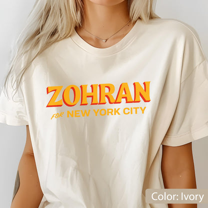 May include: Ivory-colored t-shirt with the text "ZOHRAN" in orange letters with a red outline, and "FOR NEW YORK CITY" in smaller orange letters. The shirt is a casual style.