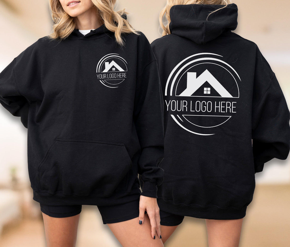 May include: Black hoodie with a white logo featuring a house silhouette and the text "YOUR LOGO HERE". The hoodie has a front pocket and a hood. The logo is also on the back of the hoodie. The model is wearing black shorts.