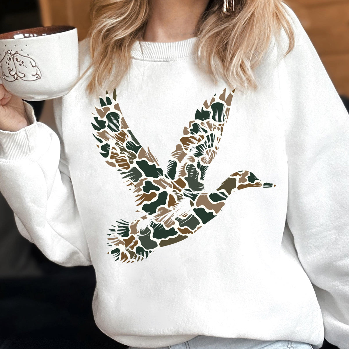 May include: A white crewneck sweatshirt featuring a large graphic of a flying duck. The duck is filled with a camouflage pattern consisting of brown, dark green, and tan organic shapes. The sweatshirt has long sleeves and a ribbed collar.