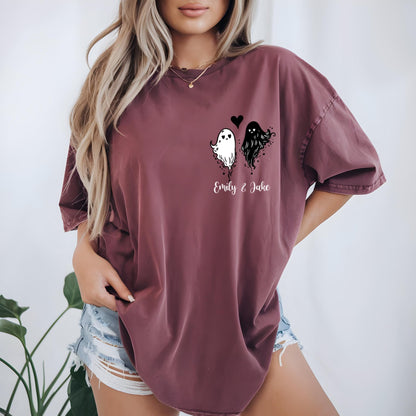May include: A maroon short-sleeve t-shirt featuring a graphic on the left chest area. The graphic displays a white ghost and a black ghost with a small heart between them, positioned above the white cursive text that reads Emily & Jake.