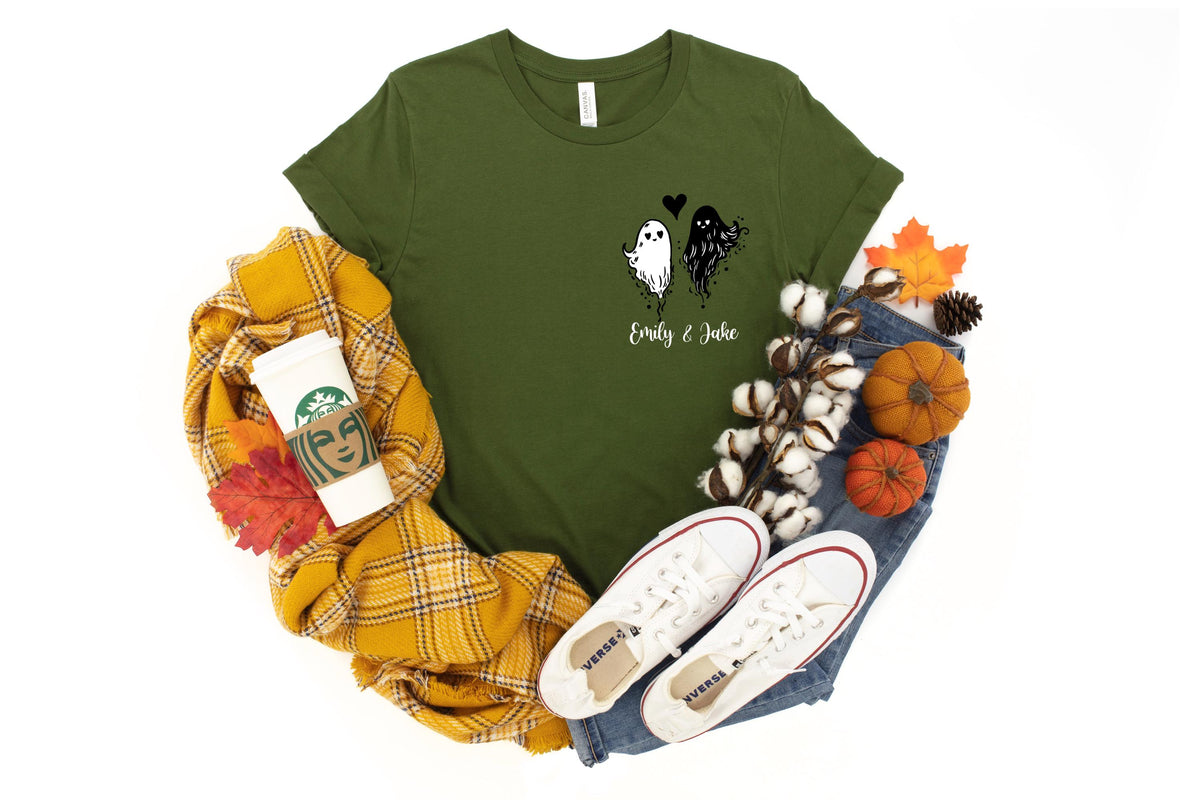 May include: An olive green t-shirt featuring a graphic of a white ghost and a black ghost with a small black heart between them. Below the ghosts, the text "Emily & Jake" is printed in a white script font. The shirt is short-sleeved with a crew neck.