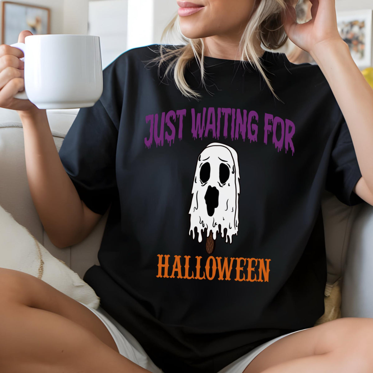 May include: Black t-shirt with the text "JUST WAITING FOR HALLOWEEN" in purple and orange. The shirt features a white ghost graphic with a sad expression. The person is holding a white mug.