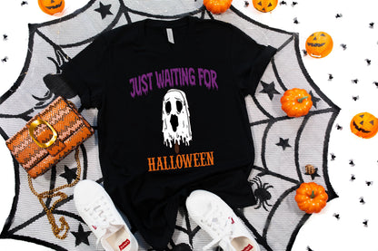 May include: Black t-shirt with the text "JUST WAITING FOR HALLOWEEN" and a white ghost graphic. The shirt is surrounded by Halloween decorations, including pumpkins, a spiderweb, a purse, and white sneakers.
