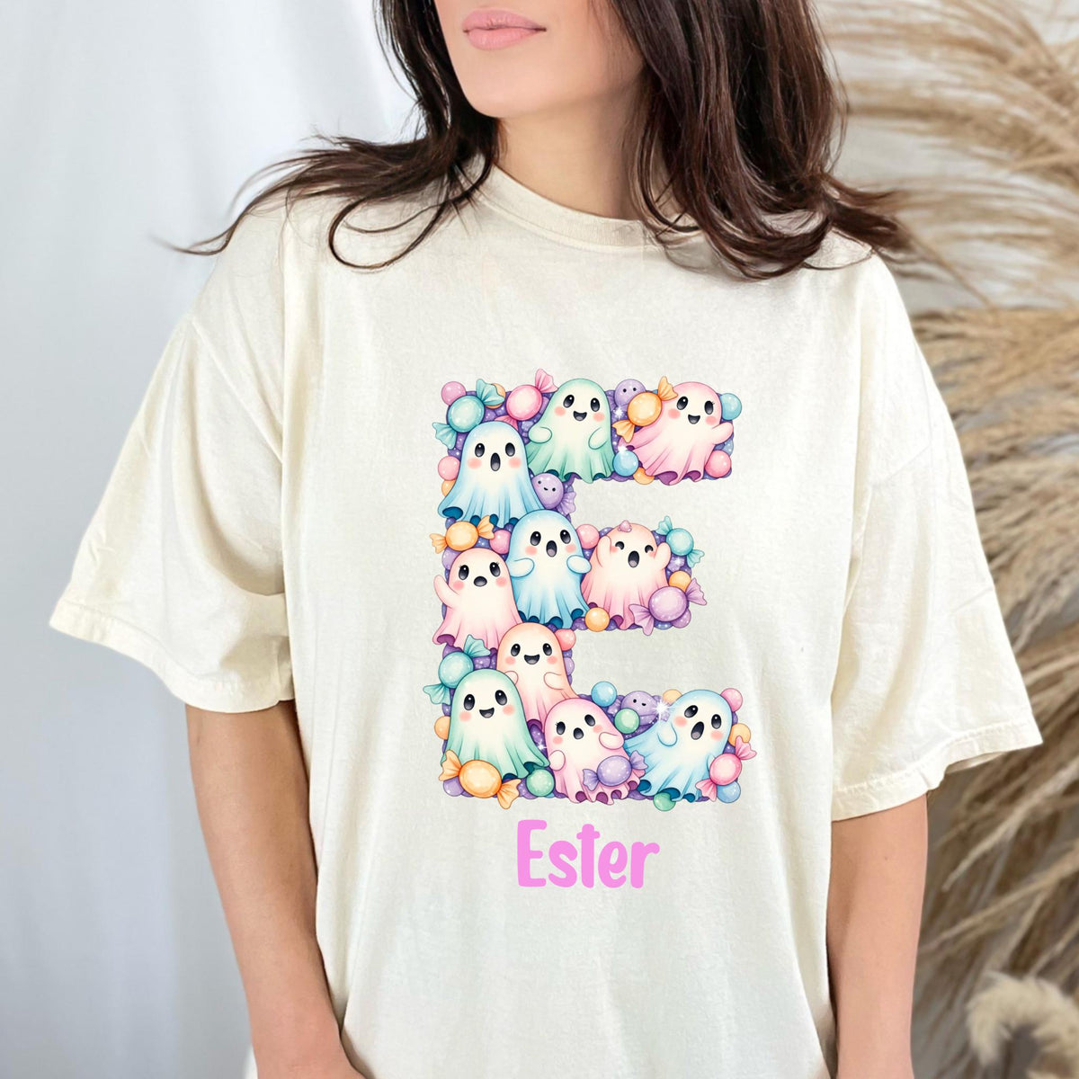 May include: A cream-colored t-shirt featuring a large letter E composed of pastel-colored cartoon ghosts and wrapped candies. Below the letter E, the name Ester is printed in a bright pink sans-serif font. The design has a soft, colorful aesthetic.