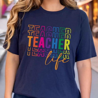 May include: Navy blue t-shirt with the words "TEACHER" in rainbow colors and the word "life" in gold script. The shirt is a crew neck and has short sleeves.