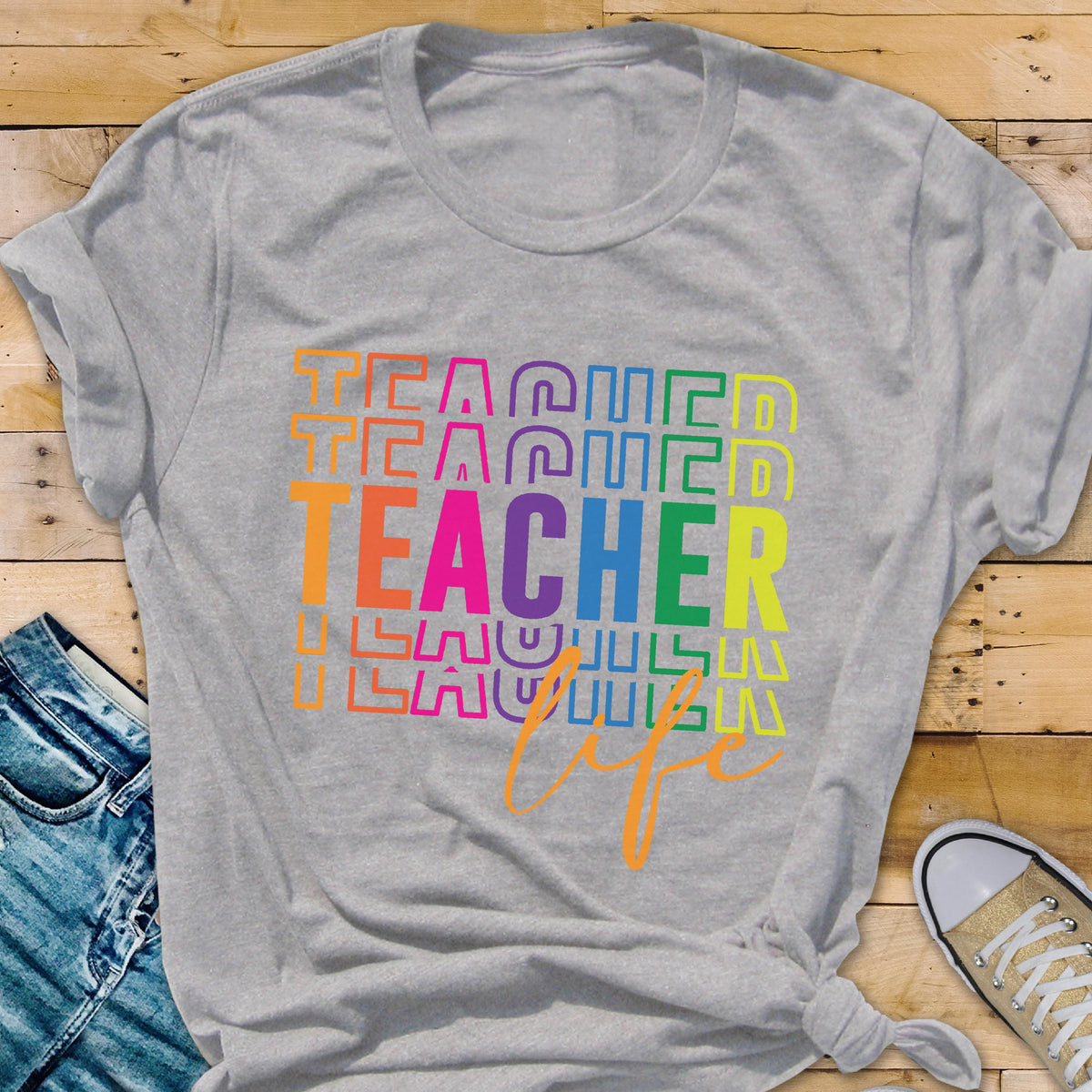 May include: A gray t-shirt with the word "TEACHER" in large, colorful letters, stacked vertically. The word "life" is written in orange script below. The shirt is paired with blue jeans and a canvas shoe.