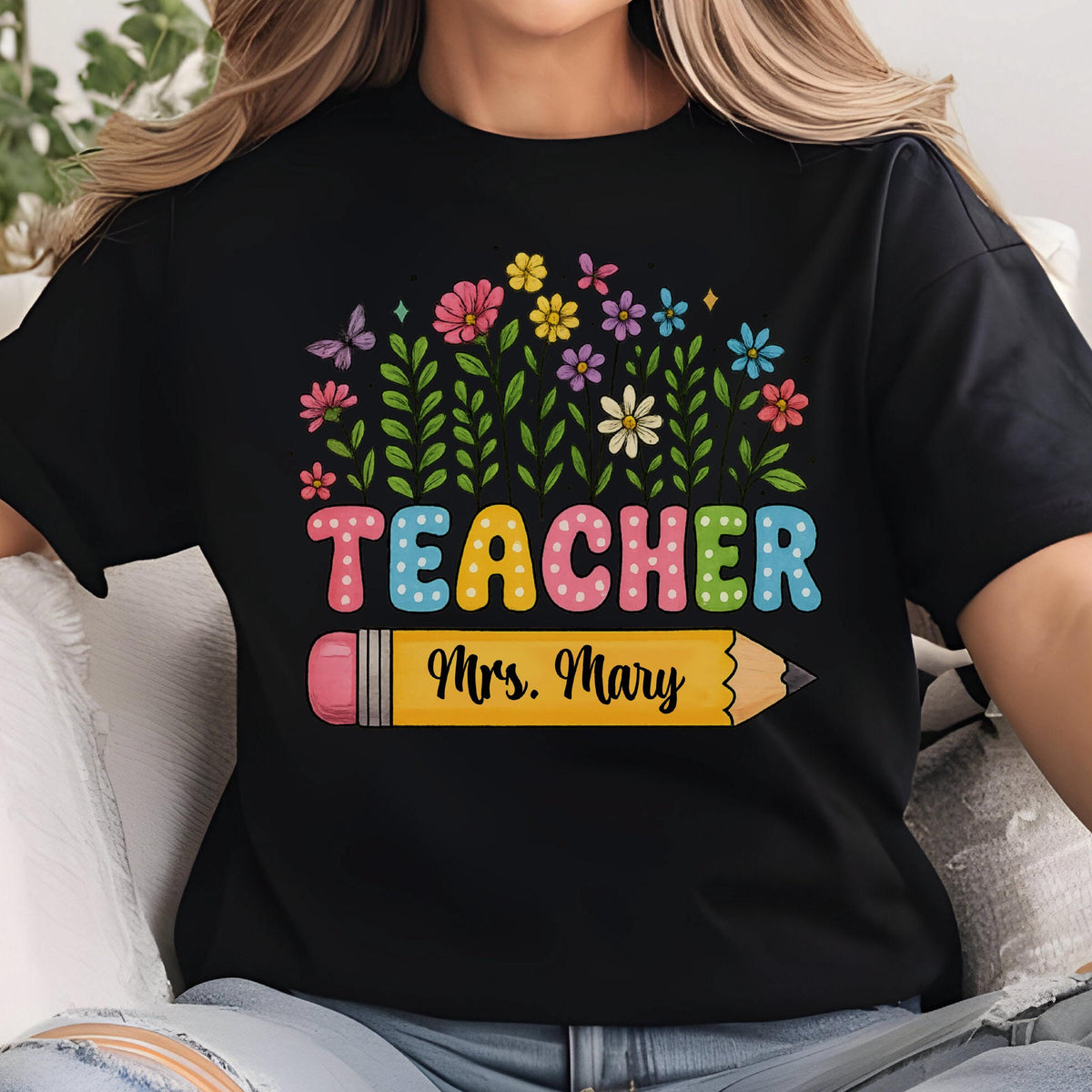 May include: Black t-shirt with the word "TEACHER" in colorful bubble letters above a yellow pencil with the name "Mrs. Mary". The design includes a floral arrangement with butterflies and various flowers.