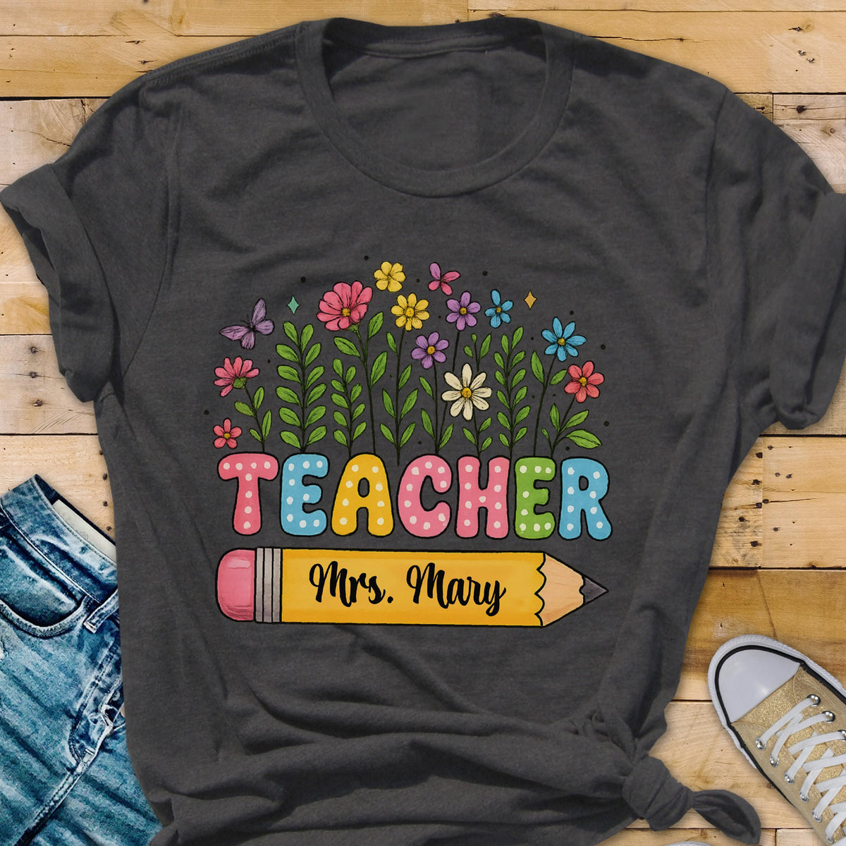 May include: Dark gray t-shirt with a colorful floral design and the word "TEACHER" in pastel letters. A yellow pencil graphic with the name "Mrs. Mary" is below the word. The shirt is a casual style.