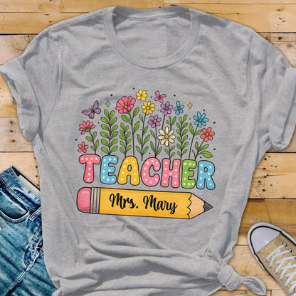 May include: A gray t-shirt featuring a colorful floral design and the word "TEACHER" in bubble letters. Below the word is a yellow pencil with the name "Mrs. Mary" printed on it. The shirt is a casual style.