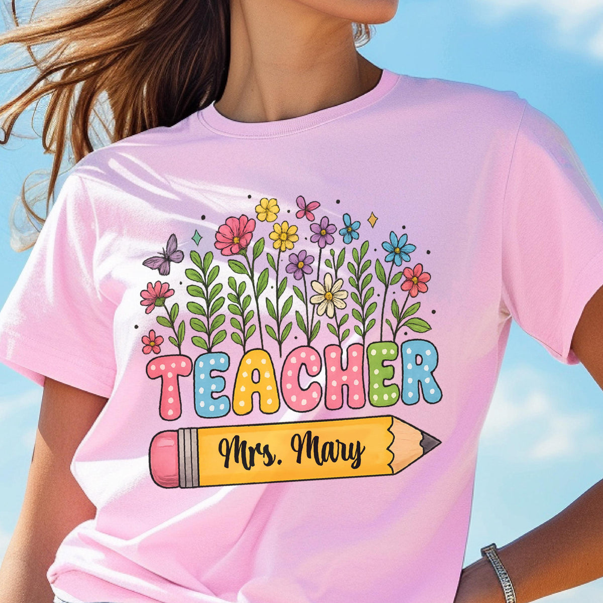 May include: Pink t-shirt with a colorful floral design and the word "TEACHER" in bubble letters. A yellow pencil graphic with "Mrs. Mary" is below the word. The shirt is a light pink color.