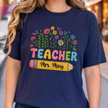 May include: Navy blue t-shirt featuring the word "TEACHER" in colorful bubble letters, with a floral design above and a yellow pencil with "Mrs. Mary" below. The design includes butterflies and various flowers.