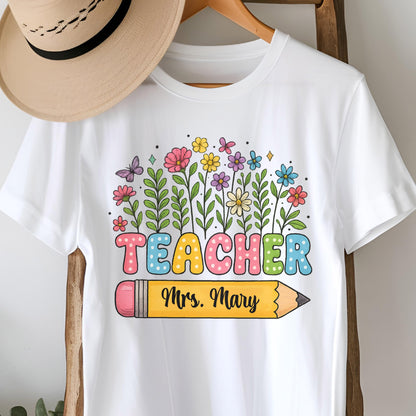 May include: White t-shirt featuring a colorful floral design with the word "TEACHER" in bubble letters and a yellow pencil with the name "Mrs. Mary". The design includes butterflies and various flowers.