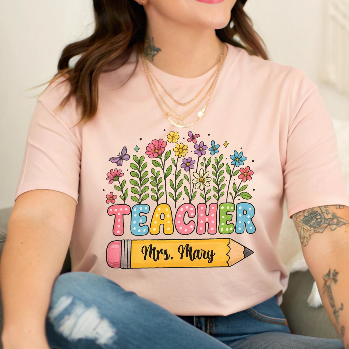 May include: A light pink t-shirt featuring a colorful floral design with the word "TEACHER" in bubble letters. Below the word is a yellow pencil with the name "Mrs. Mary". The design includes butterflies and various flowers.