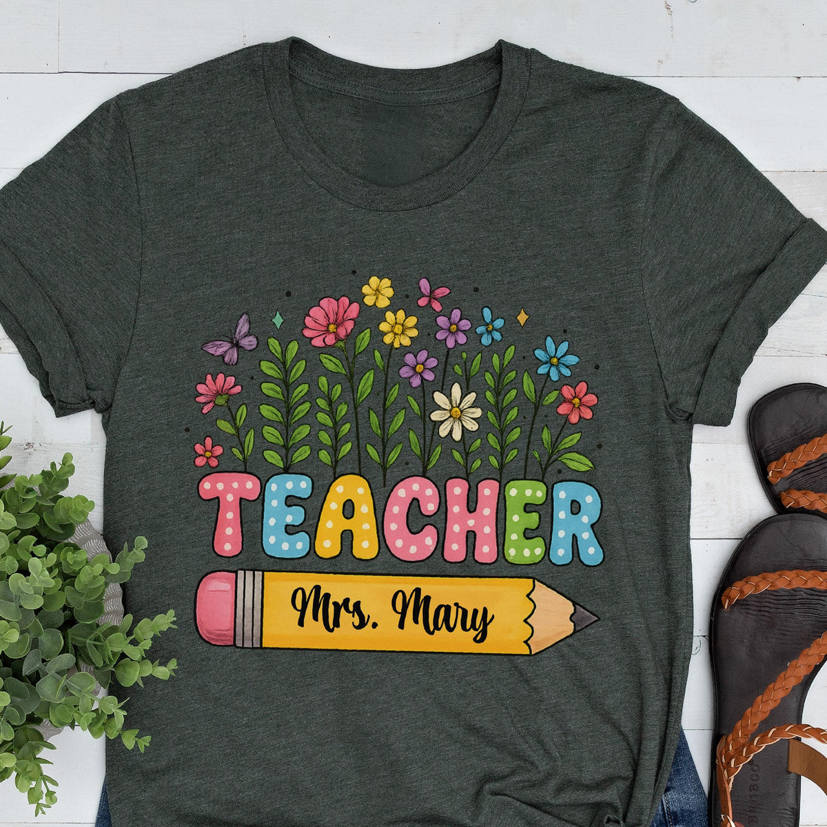 May include: Dark gray t-shirt with a colorful floral design and the word "TEACHER" in pastel letters. A yellow pencil graphic includes the name "Mrs. Mary". The shirt is a comfortable, casual style, perfect for teachers.