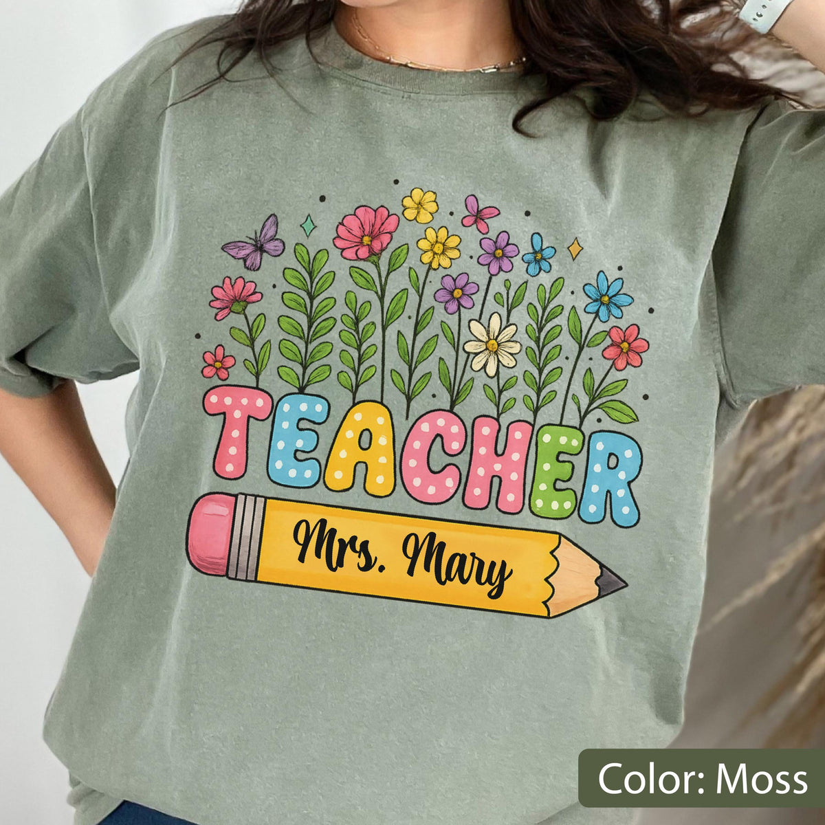 May include: Moss green t-shirt with a colorful floral design and the word "TEACHER" in bubble letters. A yellow pencil with the name "Mrs. Mary" is below the word. The shirt is a comfortable, casual style.