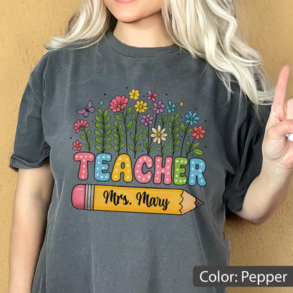 May include: Pepper-colored t-shirt featuring a colorful floral design and the word "TEACHER" in bubble letters. A yellow pencil graphic with "Mrs. Mary" is below the word. The shirt is a comfortable, casual style.