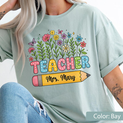 May include: A light blue t-shirt with a colorful floral design and the word "TEACHER" in bubble letters. A yellow pencil with the name "Mrs. Mary" is below the word "TEACHER". The shirt is a comfortable, casual style.
