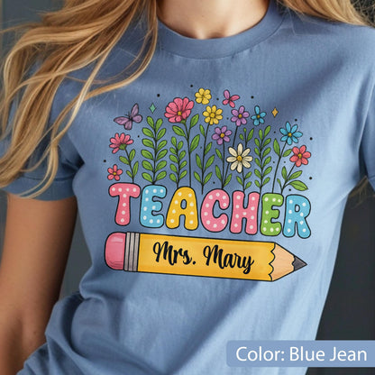 May include: Blue jean-colored t-shirt with a colorful floral design and the word "TEACHER" in bubble letters. A yellow pencil graphic with the text "Mrs. Mary" is below the word "TEACHER".