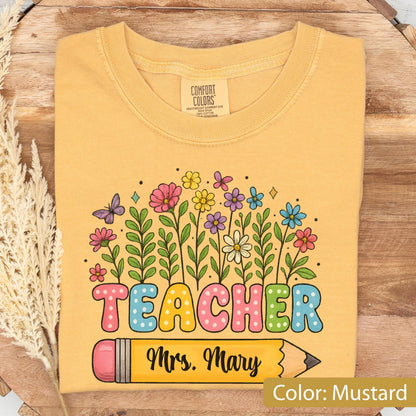 May include: Mustard-colored t-shirt with a floral design and the word "TEACHER" in colorful bubble letters. A yellow pencil graphic with "Mrs. Mary" is below the word. The shirt is made of a soft material.