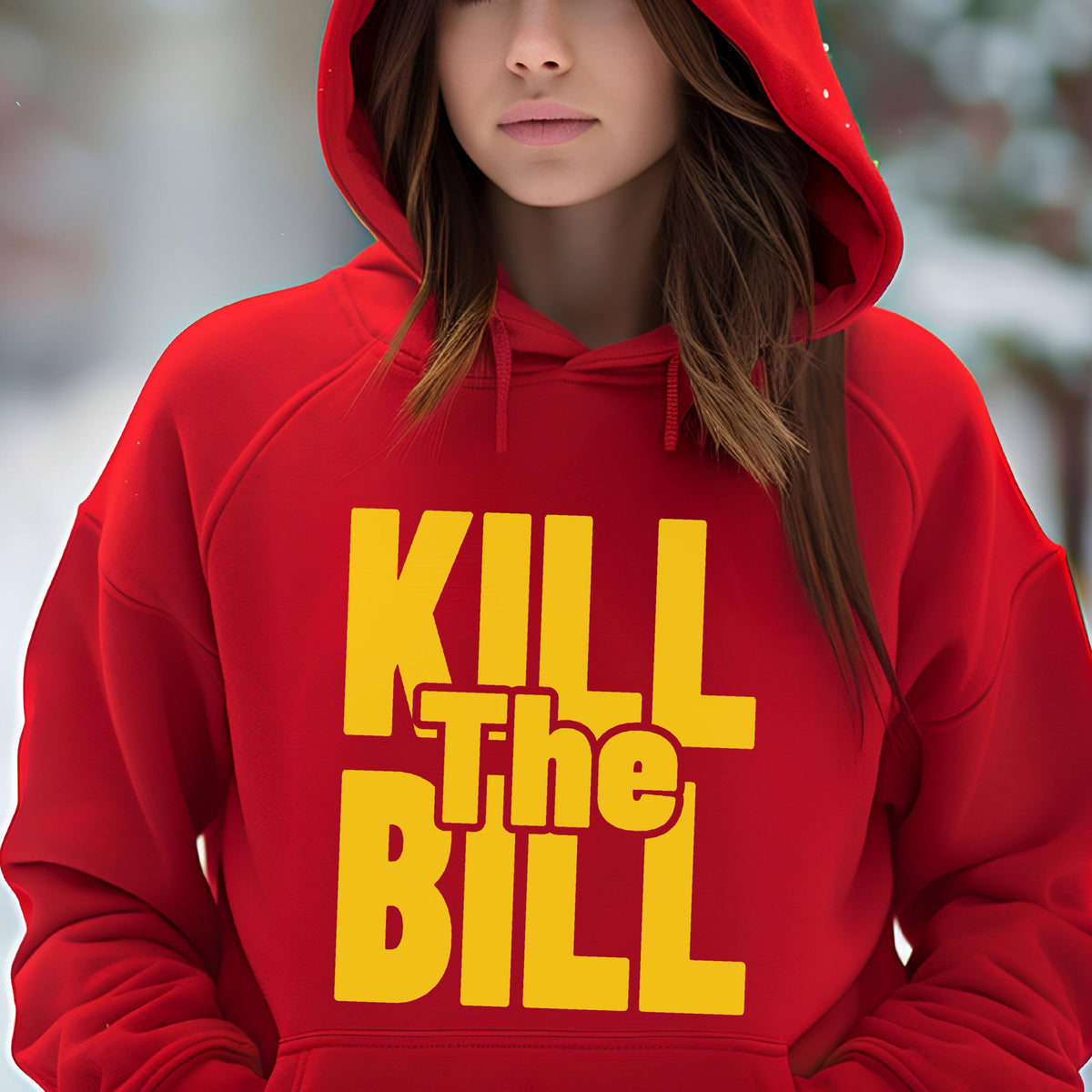 May include: A red hooded sweatshirt with the text "KILL The BILL" in yellow block letters. The sweatshirt has a drawstring hood and a front pocket. The background is blurred, suggesting an outdoor setting.