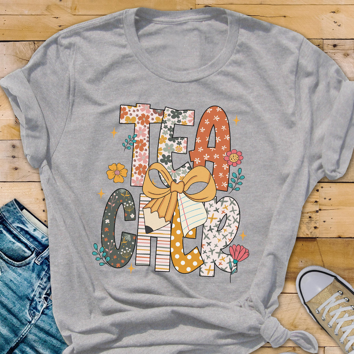 May include: A heather gray t-shirt featuring the word "TEACHER" in a colorful, floral design. The letters are adorned with flowers, patterns, and a bow, with a pencil and notepad graphic. A casual, everyday tee.