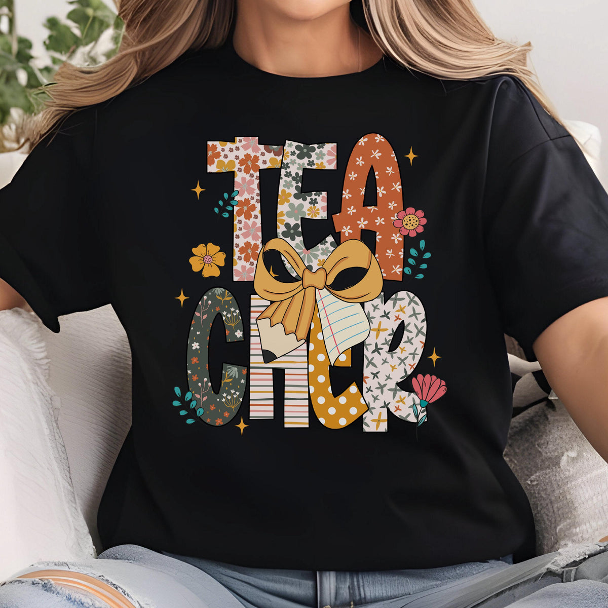May include: Black t-shirt with the word "TEACHER" in a colorful, floral design. The letters are filled with various patterns, including flowers, polka dots, and stripes. A large bow and pencil graphic are in the center.