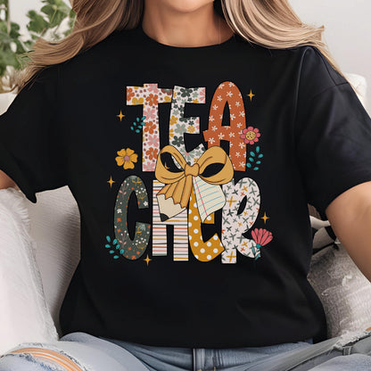 May include: Black t-shirt with the word "TEACHER" in a colorful, floral design. The letters are filled with various patterns, including flowers, polka dots, and stripes. A large bow and pencil graphic are in the center.