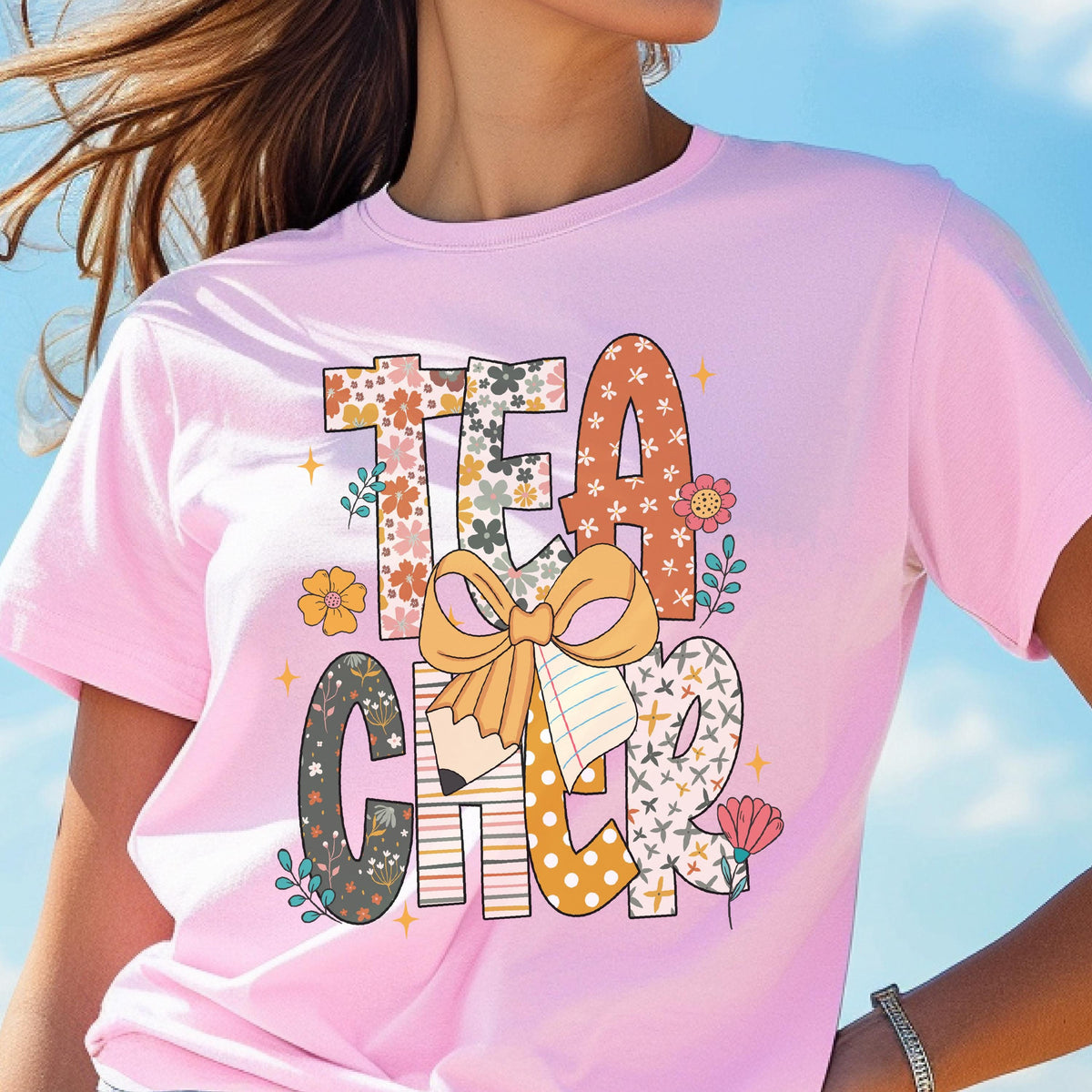 May include: Pink t-shirt with the word "TEACHER" in a colorful, floral, and patterned design. The design includes a pencil, bow, and flower accents. The shirt is a crew neck style.