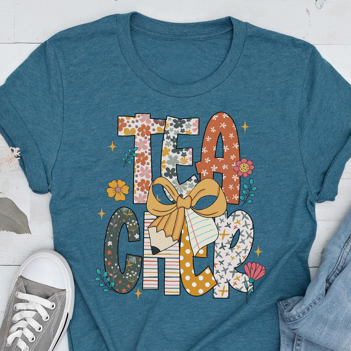 May include: Teal t-shirt with the word "TEACHER" in a colorful, floral design. The letters are decorated with flowers, polka dots, and a pencil with a bow. A gray sneaker and blue jeans are visible.