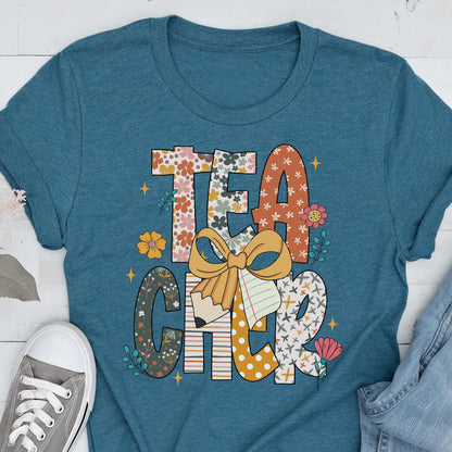 May include: Teal t-shirt with the word "TEACHER" in a colorful, floral design. The letters are decorated with flowers, polka dots, and a pencil with a bow. A gray sneaker and blue jeans are visible.