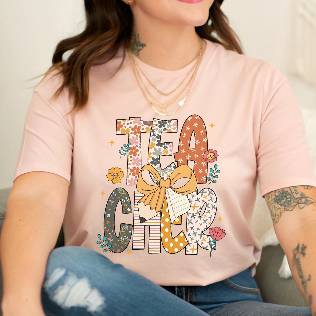 May include: A light pink t-shirt with the word "TEACHER" in a colorful, floral design. The letters are decorated with flowers, patterns, and a pencil and bow graphic. The shirt is a casual style.