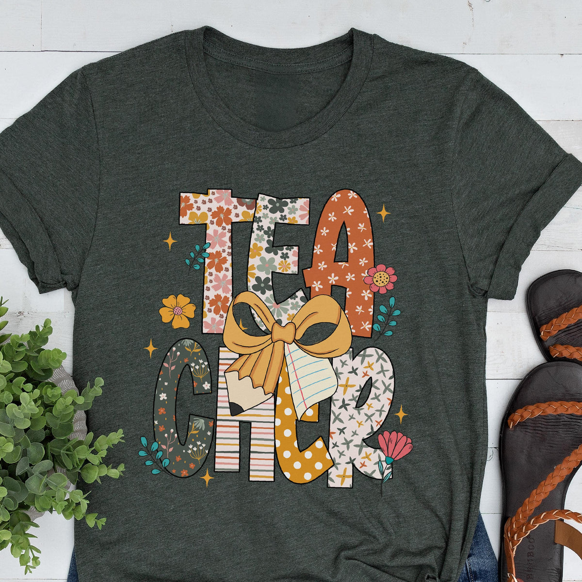 May include: Dark gray t-shirt with the word "TEACHER" in a colorful, floral design. The design includes a bow, pencil, and notebook elements. The shirt is a casual wear item.