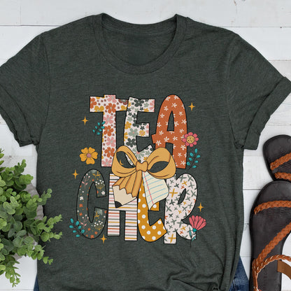 May include: Dark gray t-shirt with the word "TEACHER" in a colorful, floral design. The design includes a bow, pencil, and notebook elements. The shirt is a casual wear item.