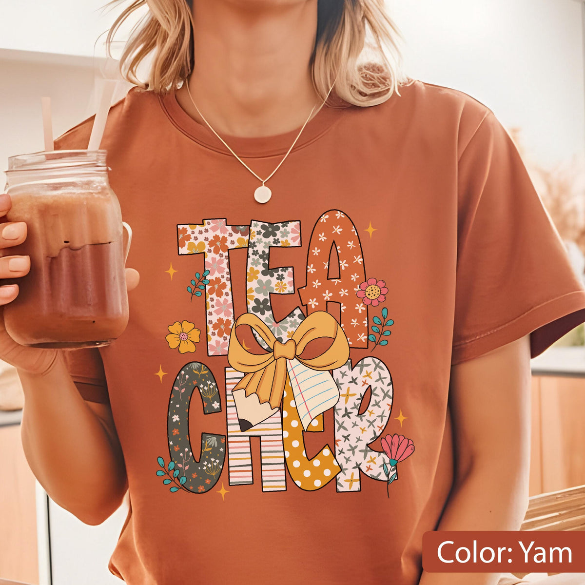 May include: A yam-colored t-shirt with the word "TEACHER" in a colorful, floral design. The design includes a pencil, a bow, and various flower illustrations. The shirt is short-sleeved and has a round neckline.