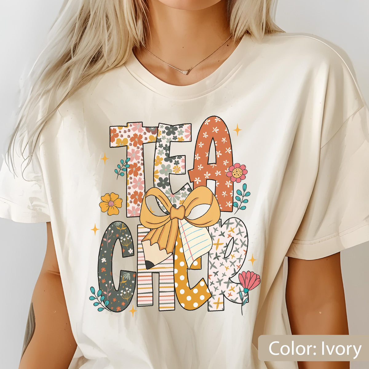 May include: Ivory-colored t-shirt with the word "TEACHER" in a colorful, floral design. The letters are decorated with patterns, flowers, and a pencil. A large bow and notebook are also featured in the design.
