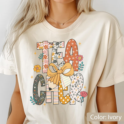 May include: Ivory-colored t-shirt with the word "TEACHER" in a colorful, floral design. The letters are decorated with patterns, flowers, and a pencil. A large bow and notebook are also featured in the design.