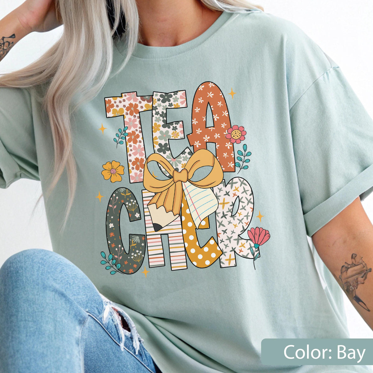 May include: A light blue t-shirt with the word "TEACHER" in a colorful floral design. The letters are filled with various patterns and flowers, with a bow and pencil graphic in the center. The shirt is a relaxed fit.